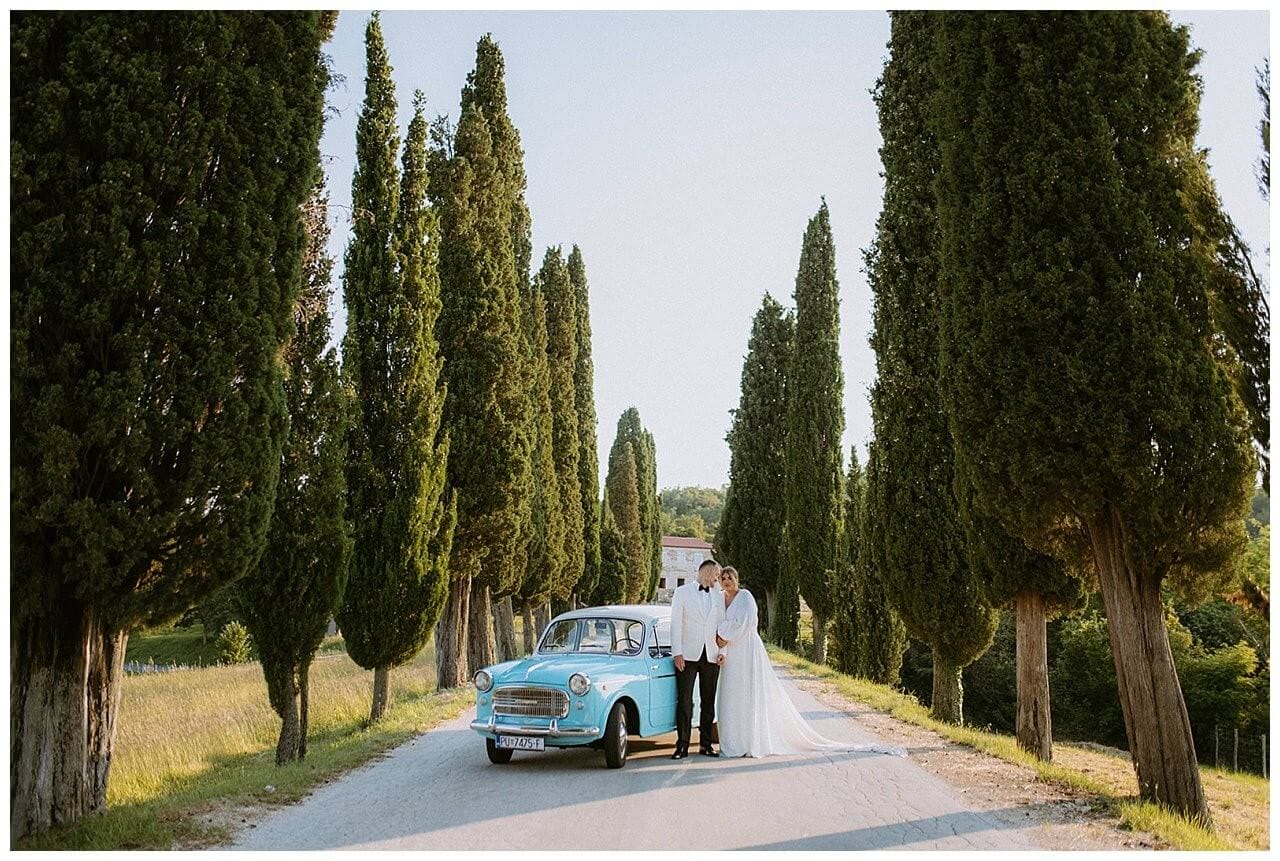 Brautpaar in weiß und einen Türkisblauen Oldtimer auf einen abgelegenen Gehweg mit Bäumen bei ihrer Hochzeit in Istrien Kroatien